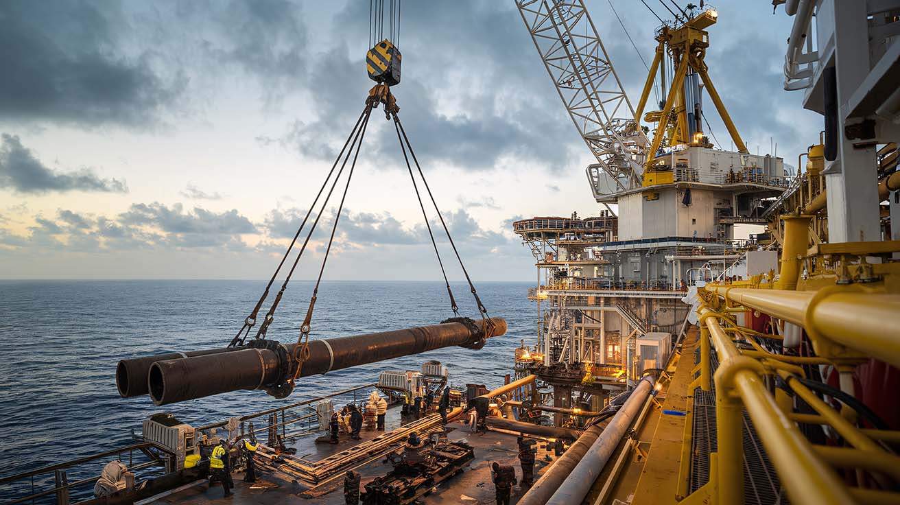 Offshore pipe installation at sunset, with large steel pipes being lifted by cranes and secured with pipe lifting tools.